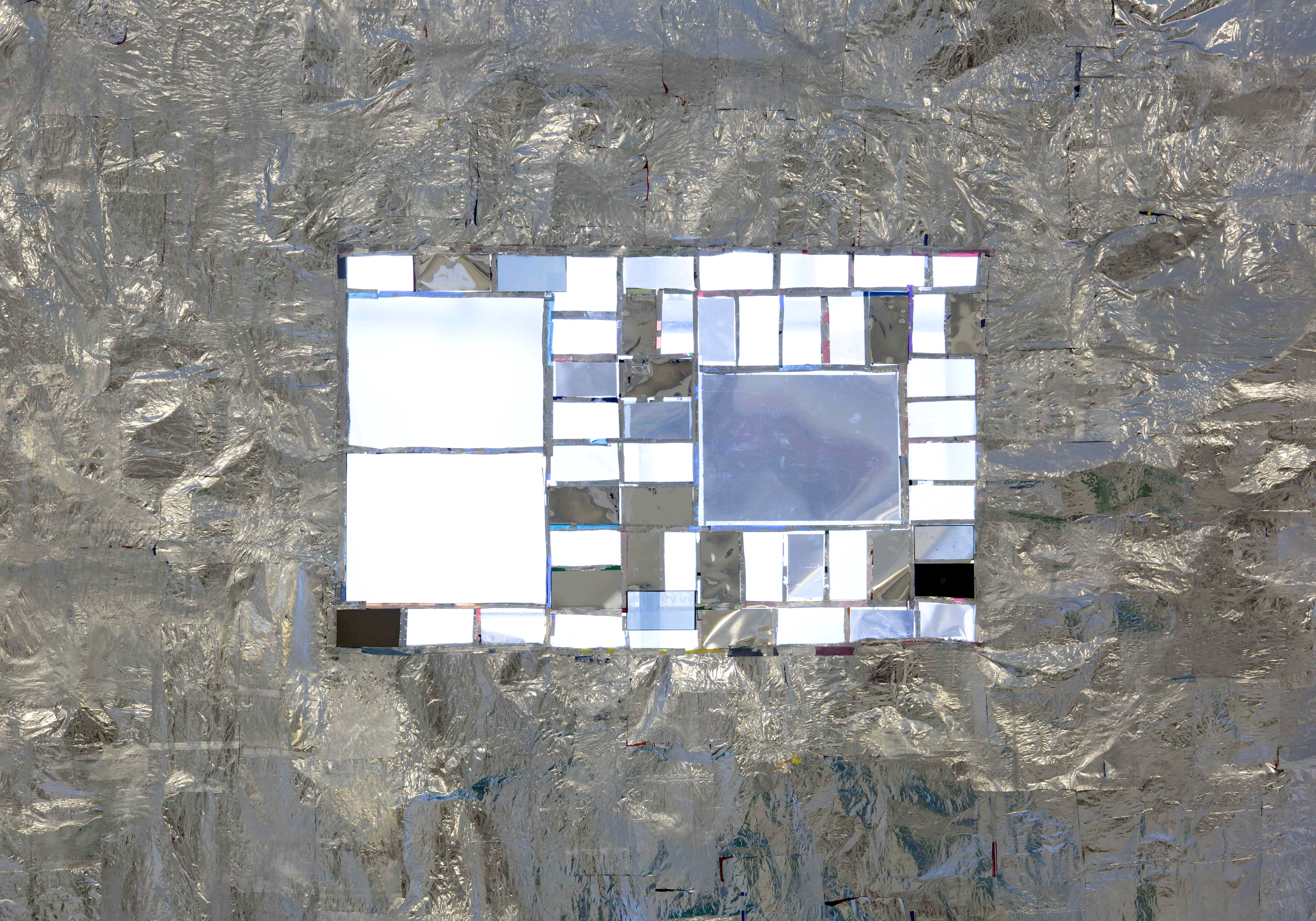 An image showing a rectangular sheet made from crisp packets, ironed together with a crinkly surface. In the centre is a landscape format rectangle made up of smaller and larger rectangular, semi-transparent shapes which are made from mobile phone and laptop screen parts